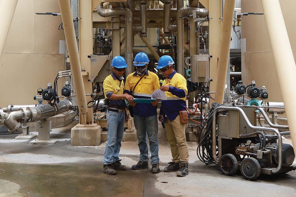 Three Thai men in safety gear stand, jointly consulting a clipboard. In the background is a tangle of pipes and industrial machinery.