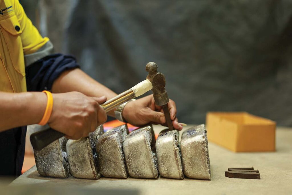 A woman uses a hammer and die to make markings on a row of silver-grey metallic ingots.