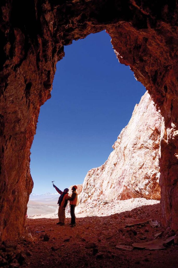 Two men in orange safety vests are framed against a blue sky by a cave opening. One man is pointing at the cave wall with a geologists' hammer while the other makes notes.